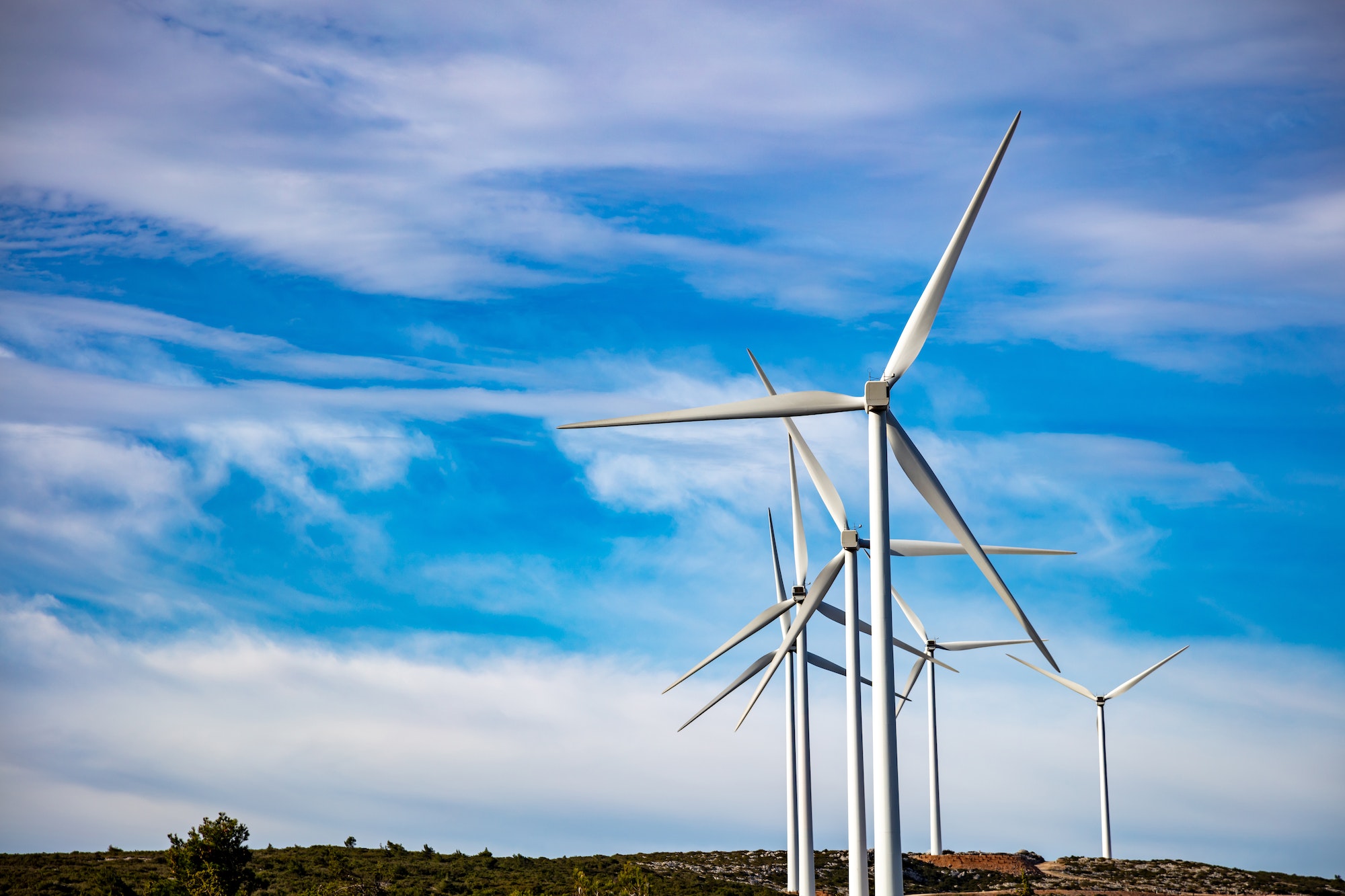 A row of wind turbines sitting on a rocky hill with a background of blue sky and wispy clouds